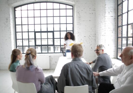 Vrouw met donker krulhaar staat recht en spreekt groepje mensen toe die neerzitten op een witte stoel in een witgekleurde ruimte