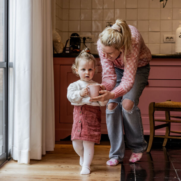 jonge vrouw helpt meisjespeuter kopje koffie dragen. Ze zijn gekleed in het roze en stappen in een roze keuken