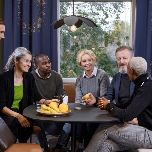 6 vrienden van verschillende leeftijd voeren een gesprek rond een ronde tafel met in het midden een toestel dat het geluid versterkt.