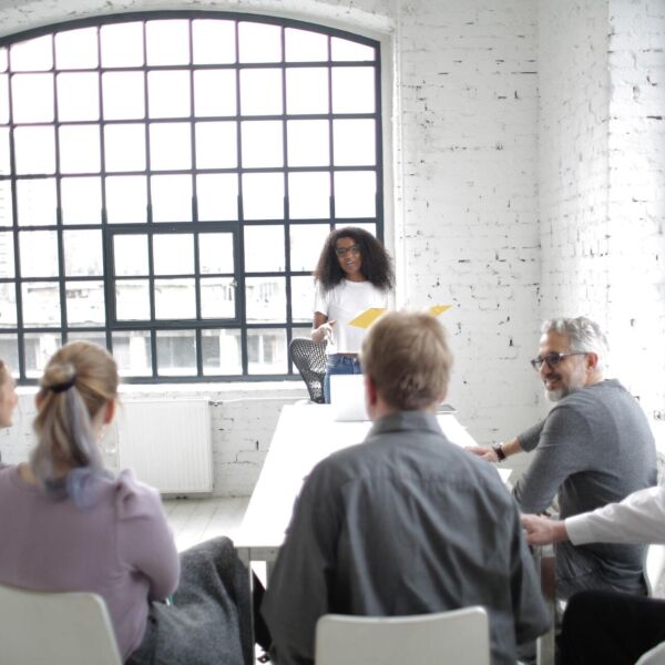 Vrouw met donker krulhaar staat recht en spreekt groepje mensen toe die neerzitten op een witte stoel in een witgekleurde ruimte
