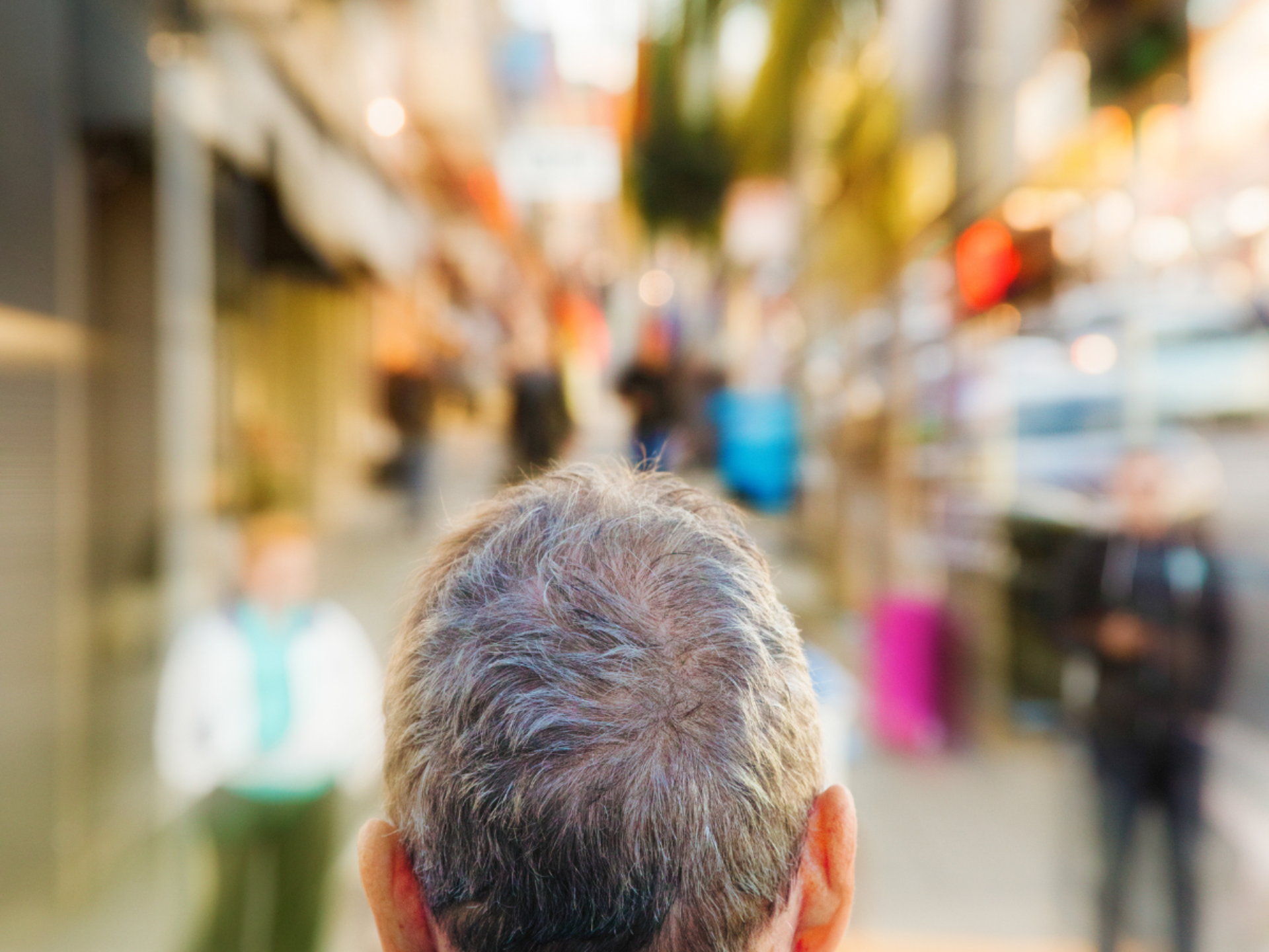 Man met kort grijs haar loopt in een drukke winkelstraat. De achtergrond is wazig omdat hij duizelig wordt van alle prikkels.